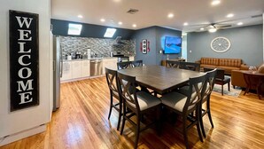 A modern kitchen with a built-in oven, a dining area with a wooden table and chairs, a living room with a sofa and a TV, and a 'WELCOME' sign on the wall. - MODERN MASSIVE LARGE GROUP 3800sf MIT HARVARD BOS (Somerville)