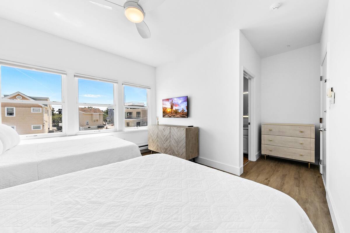 Un dormitorio con dos camas, un mueble con diseño tipo espiga, un cuadro y vistas de edificios a través de amplios ventanales.