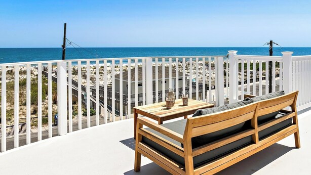 A wooden bench and table on a balcony with a white railing, overlooking a beach with a clear blue sky. - The Hideaway Hotel (Beach Haven)