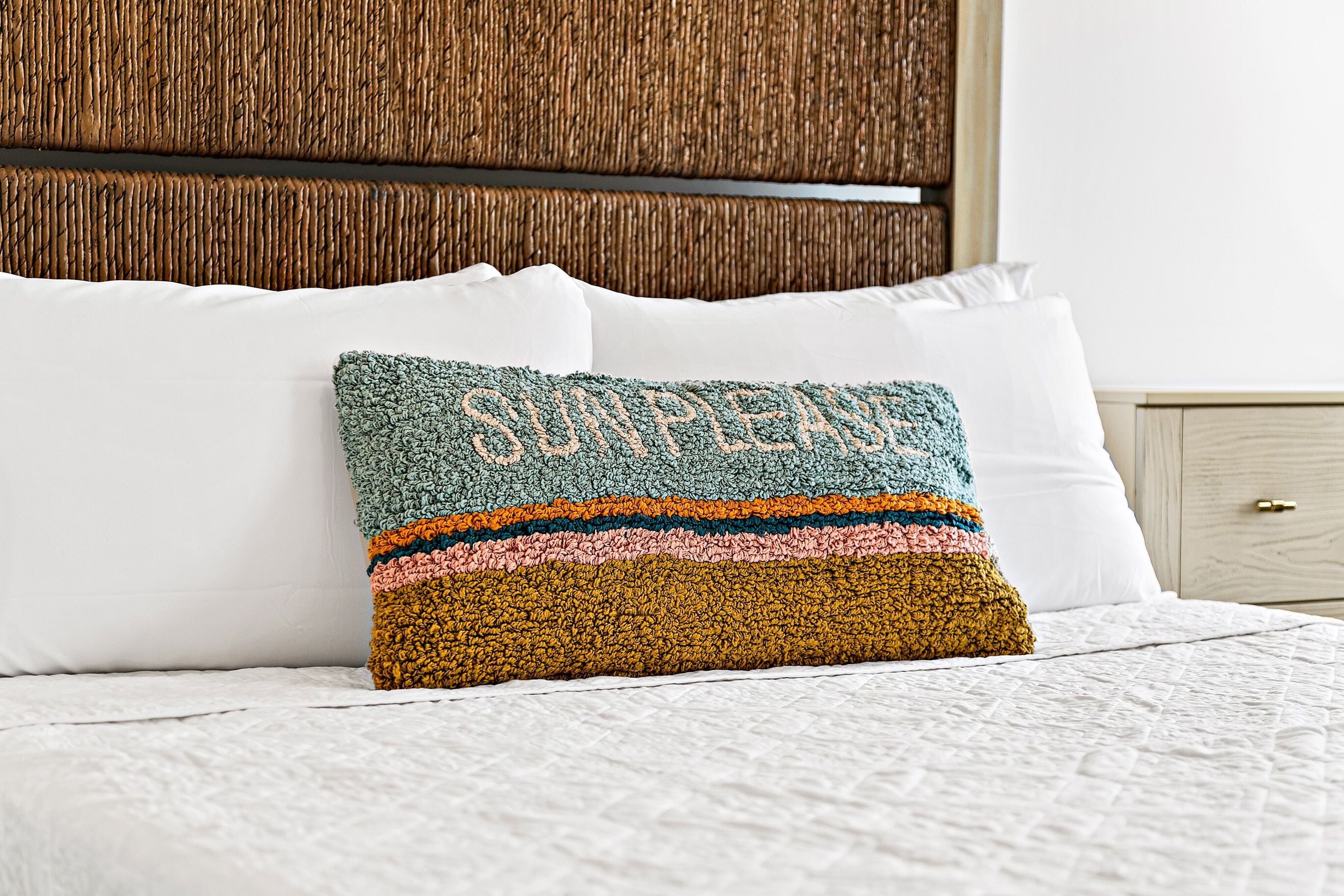 A bed with white linens, a colorful knitted pillow, and a wooden headboard.