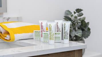 Una encimera de baño con artículos de aseo, incluyendo una toalla amarilla y blanca, y varias botellas de productos para el cuidado corporal Malin+Goetz.