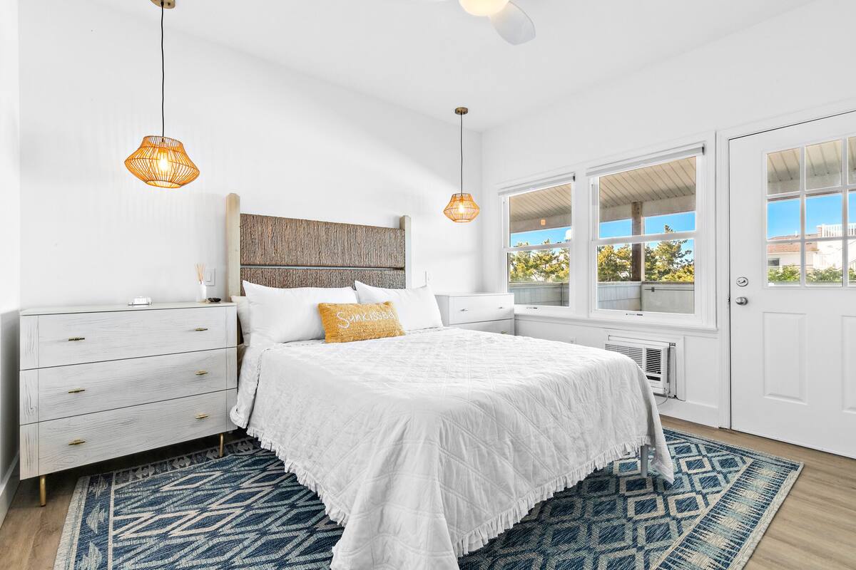 Un dormitorio con cama, cómoda blanca, dos lámparas colgantes, una ventana y una alfombra estampada.
