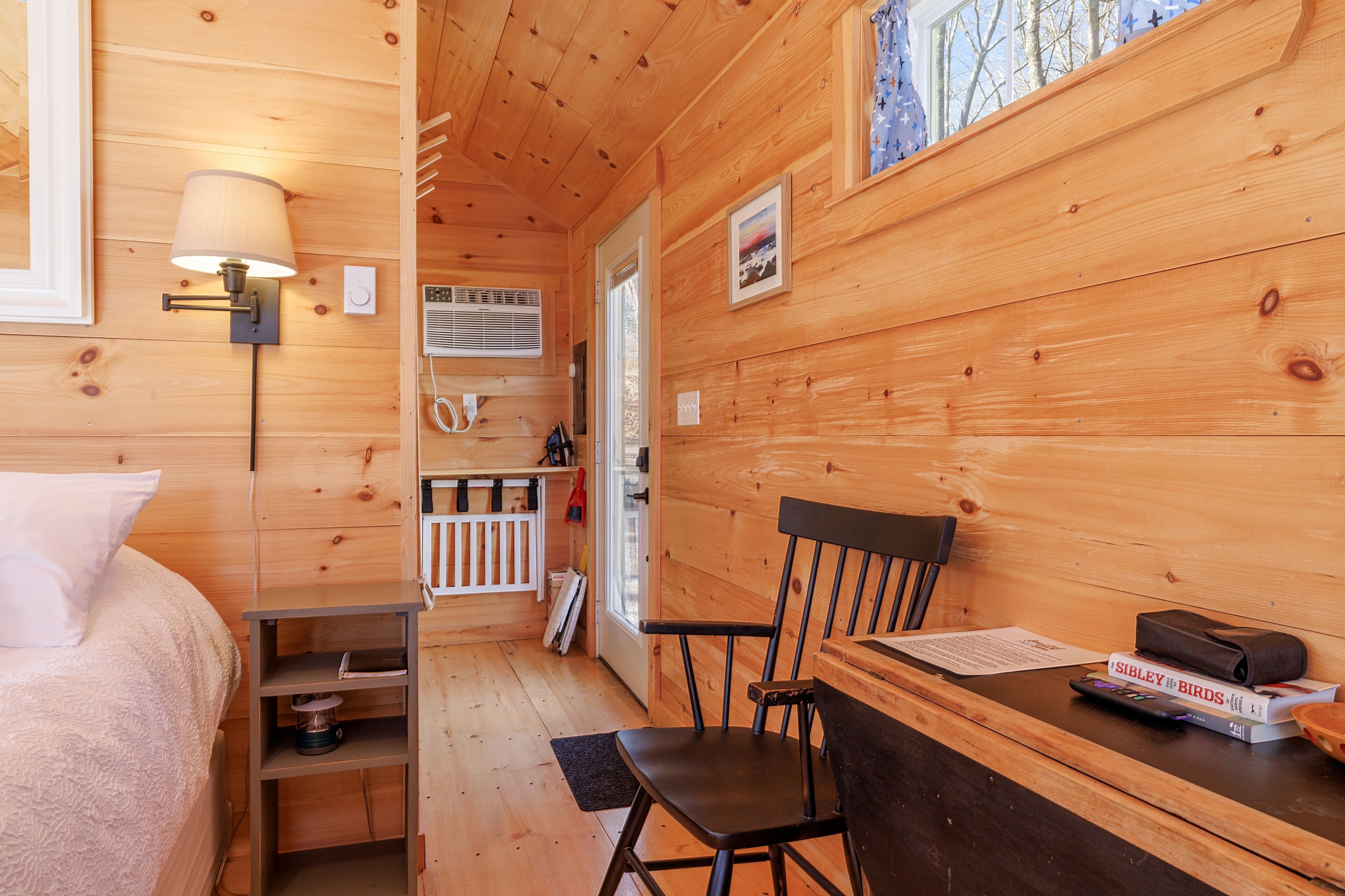 Una habitación tipo cabaña de madera con una cama, un escritorio con una silla, una lámpara y una ventana con vistas a árboles.