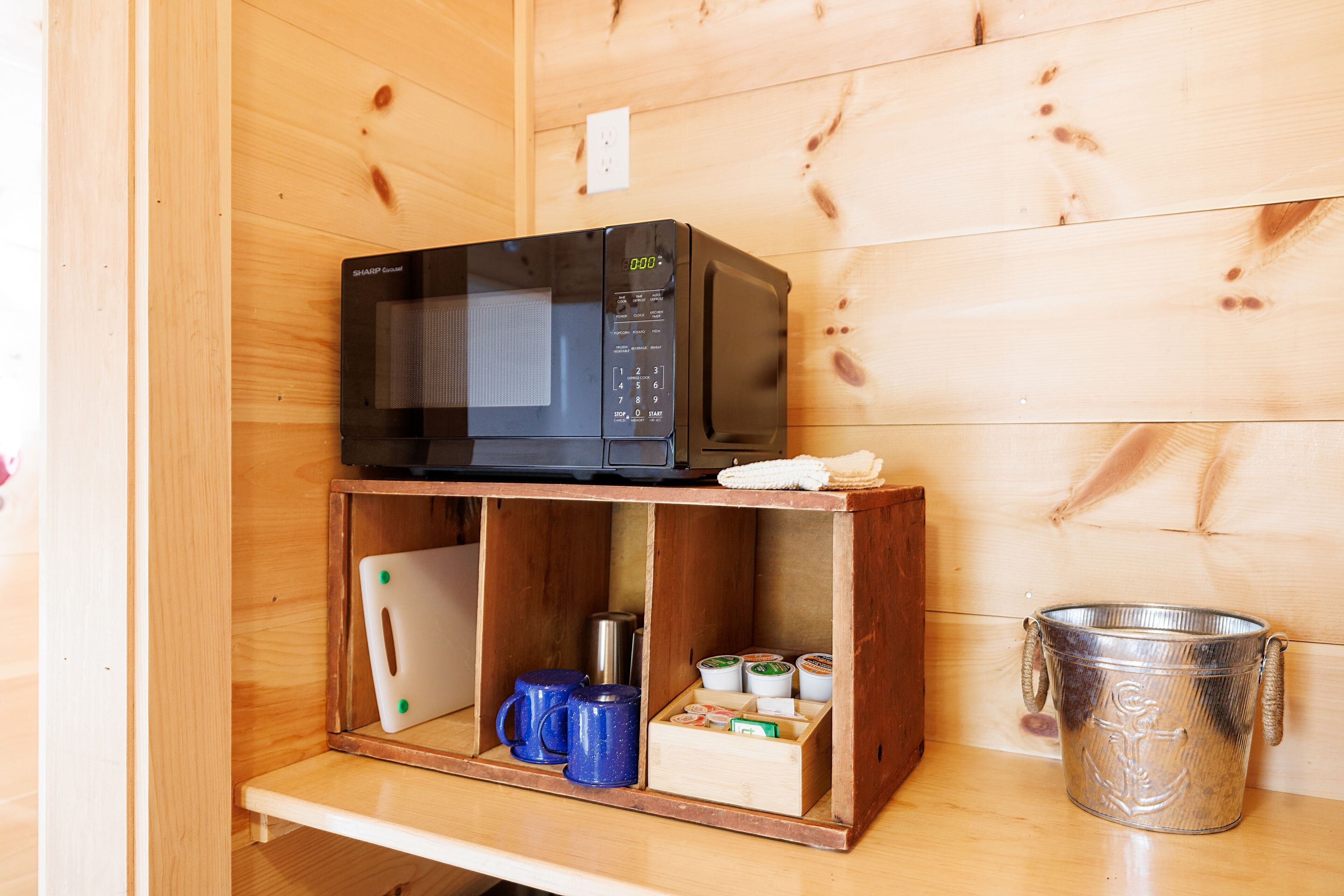 Un mueble de madera con un microondas encima, un pequeño estante con tazas, una caja y un recipiente metálico.