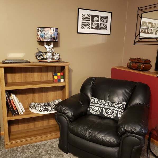 A leather armchair with a patterned pillow, a wooden bookshelf with books and a Rubik's cube, a lamp, and framed artwork on the wall.
