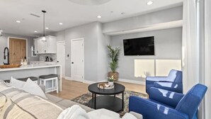 A modern living room with a sofa, a coffee table, and a blue armchair. There is a kitchen area with white cabinets and a dining table. A flat-screen TV is mounted on the wall. - Entire Brand New Luxury Townhouse! #2 (San Diego)