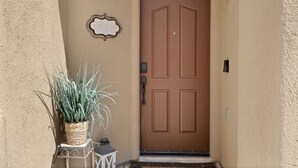 Pared beige con una puerta marrón, un cartel de bienvenida y una planta en maceta.
