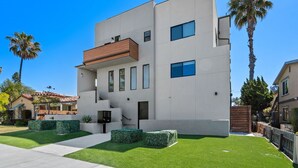 A modern two-story building with a flat roof, a wooden balcony on the second floor, and a well-maintained lawn with hedges. - Entire Brand New 2-Bedroom #5 Luxury Townhouse! (San Diego)