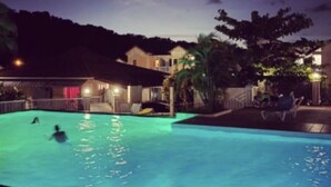 Une piscine éclairée la nuit, avec des personnes dans l’eau, entourée d’une maison et d’arbres.