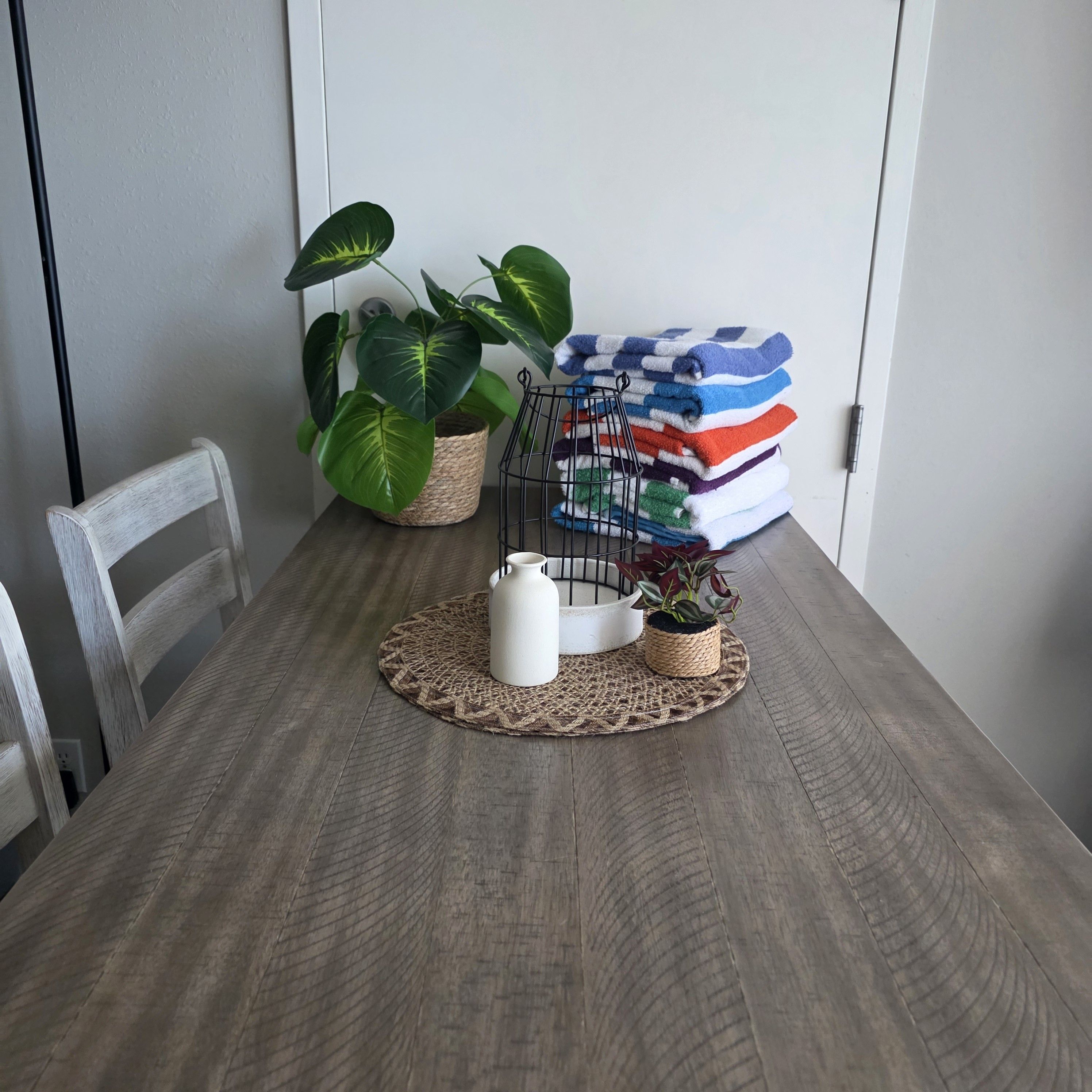 Een eettafel met een geweven placemat, een witte kruik, een kleine potplant, een metalen plantenstandaard en een stapel opgevouwen handdoeken.