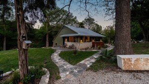 A rustic cabin with a stone pathway leading to the entrance, surrounded by trees and a well-maintained lawn. - River Run - Luxurious Adults Only Riverfront Oasis (Wimberley)