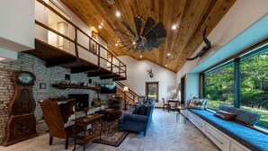 A spacious living room with a stone fireplace, wooden beams, a large window with a view of greenery, and a blue sofa. - River Run - Luxurious Adults Only Riverfront Oasis (Wimberley)