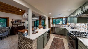 A modern kitchen with a stone island, green cabinets, and a large window offering a view of trees. - River Run - Luxurious Adults Only Riverfront Oasis (Wimberley)