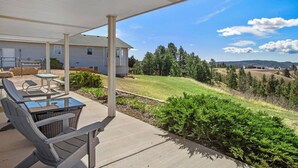 A covered patio with outdoor furniture, a view of a house, and a grassy landscape with trees. - Unique Barn Retreat by the Hills (Rapid City)