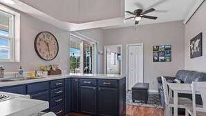 A modern kitchen with dark blue cabinets, a large clock, and a dining area with a gray couch and white chairs. - Unique Barn Retreat by the Hills (Rapid City)
