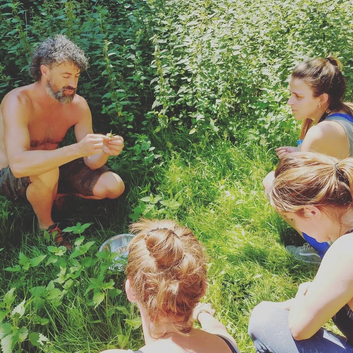 A group of people sitting outdoors in a grassy area with dense foliage in the background.