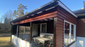 Een overdekt terras met een tafel, stoelen, een raam en een deur naar het huis.