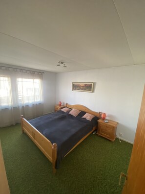 A bedroom with a wooden bed, green carpet, a nightstand with a lamp, and a painting on the wall.