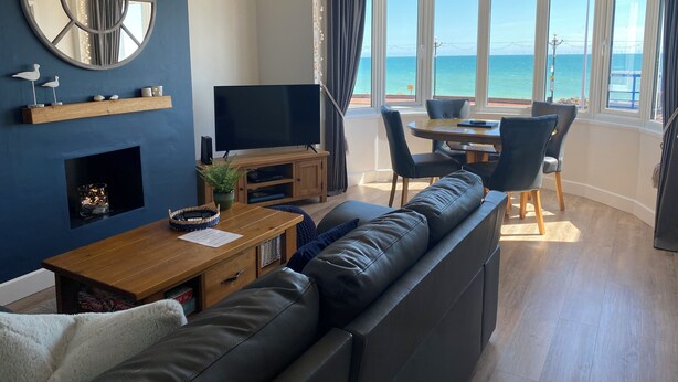 A modern living room with a large TV, comfortable seating, a fireplace, and a view of the sea. - Beach 180: Beachfront Apartment, Sea views & Private Driveway Parking  (Worthing)