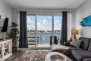 A modern living room with a sofa, a television, a plant, and a view of a body of water through large sliding doors.