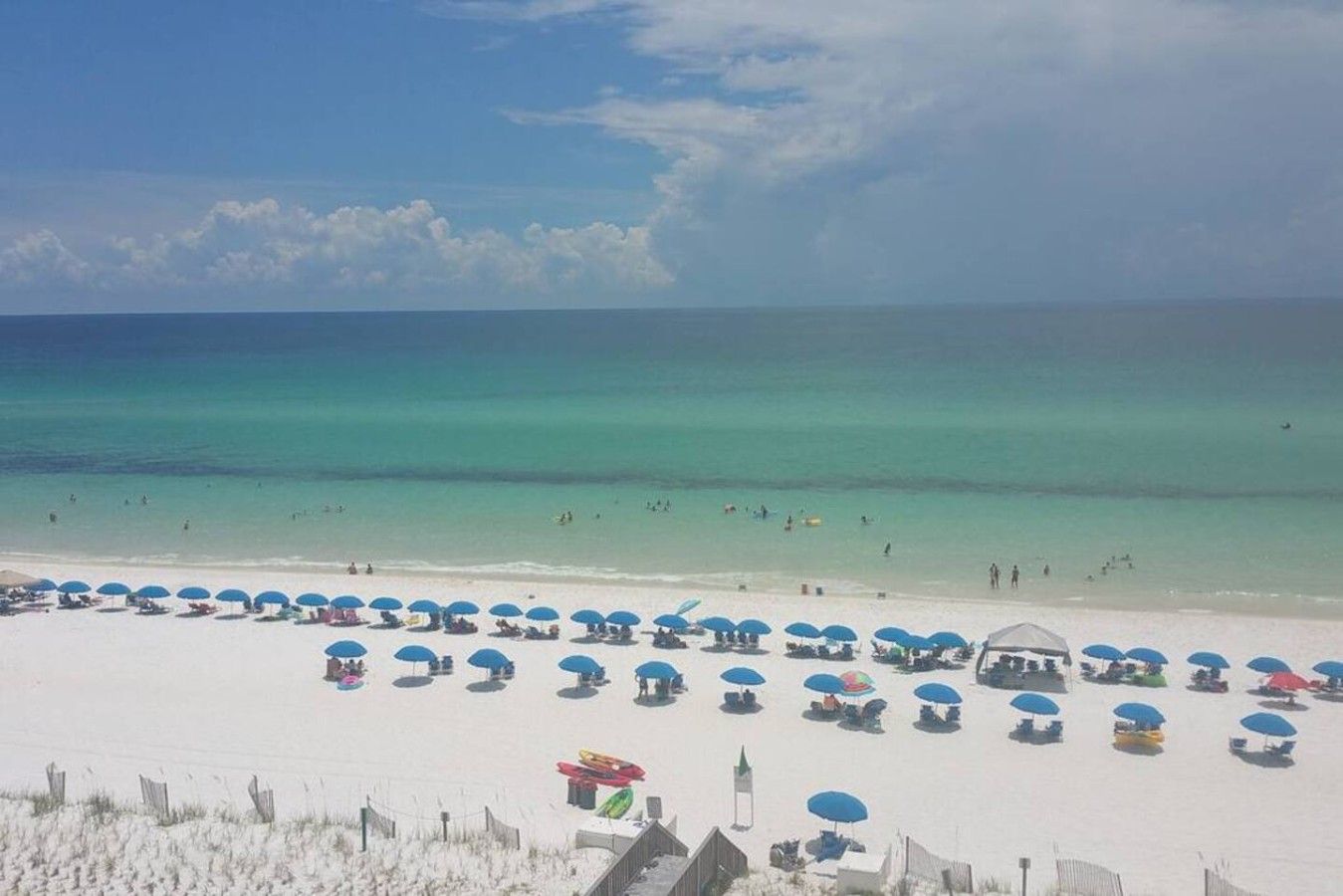 A beach with white sand, clear turquoise water, and numerous blue umbrellas.