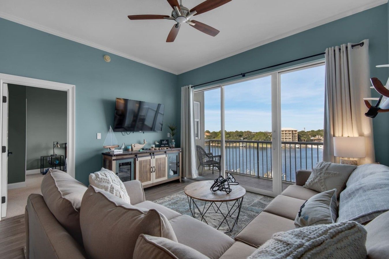 A modern living room with a large sofa, a coffee table, and a television. There is a balcony with a view of the water.