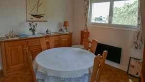 Ein Essbereich mit einem runden Tisch, der mit einer weißen Spitzenleinentischdecke bedeckt ist, Holzstühle, eine Holzkommode, ein kleiner Tisch mit einer Lampe und einer Segelboot-Dekoration an der Wand.