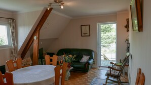 Ein gemütliches Wohnzimmer mit einem grünen Sofa, einem Holzesstisch und Stühlen. Eine Treppe führt zu einer oberen Ebene.