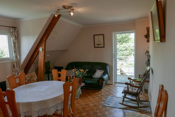 A cozy living room with a green sofa, wooden dining table, and chairs. A staircase leads to an upper level.