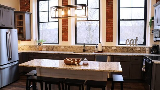 A modern kitchen with granite countertops, a central island with bar stools, a stainless steel refrigerator, and a brick accent wall. - Lake Lookout Penthouse Loft (Boyne City)