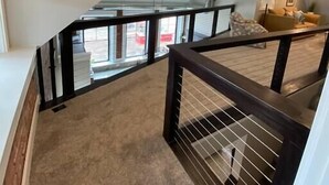 A modern loft-style apartment with a spacious living area, a staircase with a glass railing, and a view of the outdoors. - Lake Lookout Penthouse Loft (Boyne City)