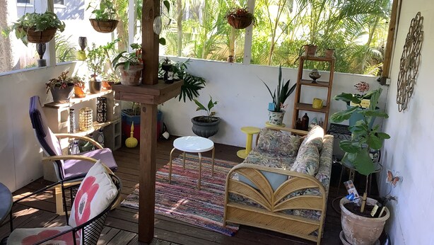 Une terrasse couverte avec un canapé en osier, une chaise et une petite table. On y trouve des plantes en pot et une étagère avec des objets décoratifs.