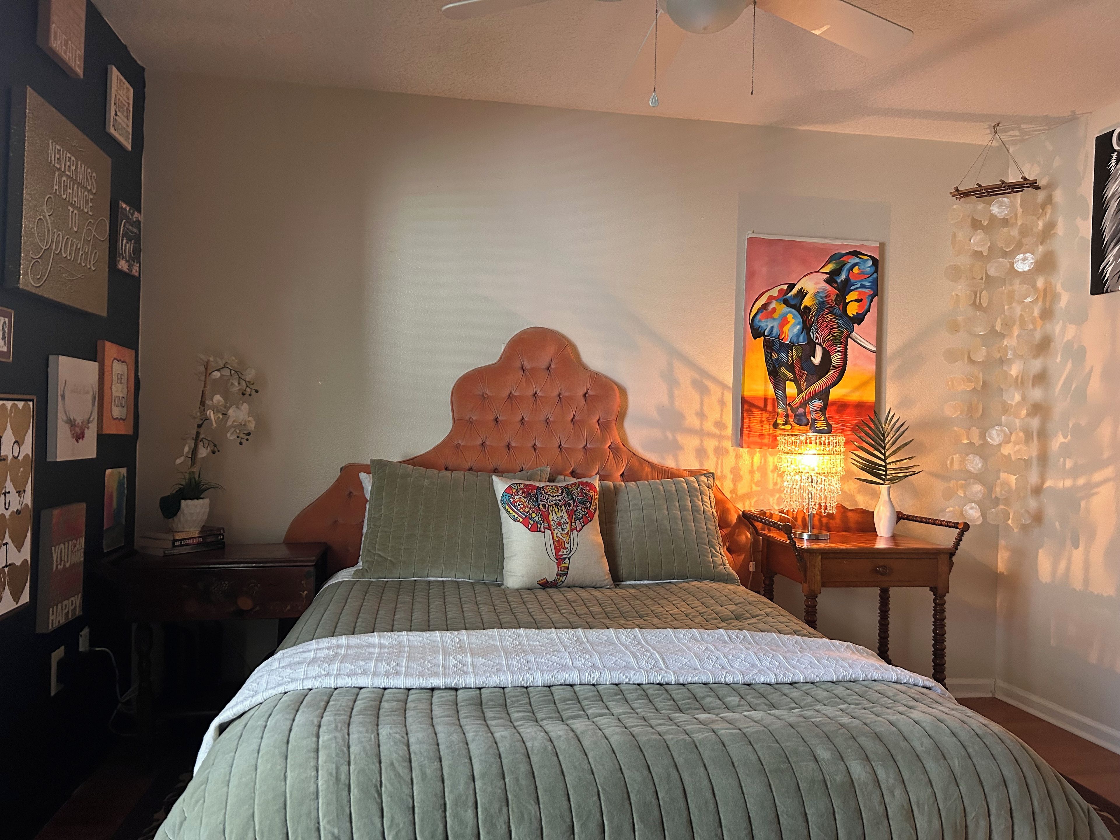 Un dormitorio con cabecera capitonada, cama con colcha verde, mesita de noche con lámpara y una pared con obras de arte enmarcadas.