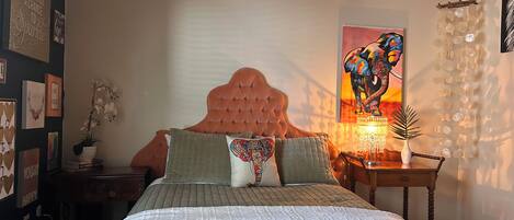 Un dormitorio con cabecera capitonada, cama con colcha verde, mesita de noche con lámpara y una pared con obras de arte enmarcadas.
