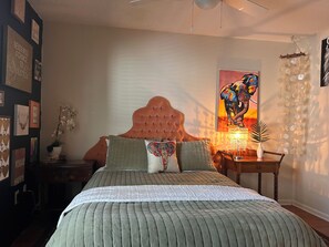 A bedroom with a tufted headboard, a bed with a green quilt, a nightstand with a lamp, and a wall with framed artwork.