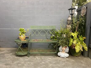 Un banc en métal vert adossé à un mur gris, entouré de plantes en pot et agrémenté d’une lampe décorative.