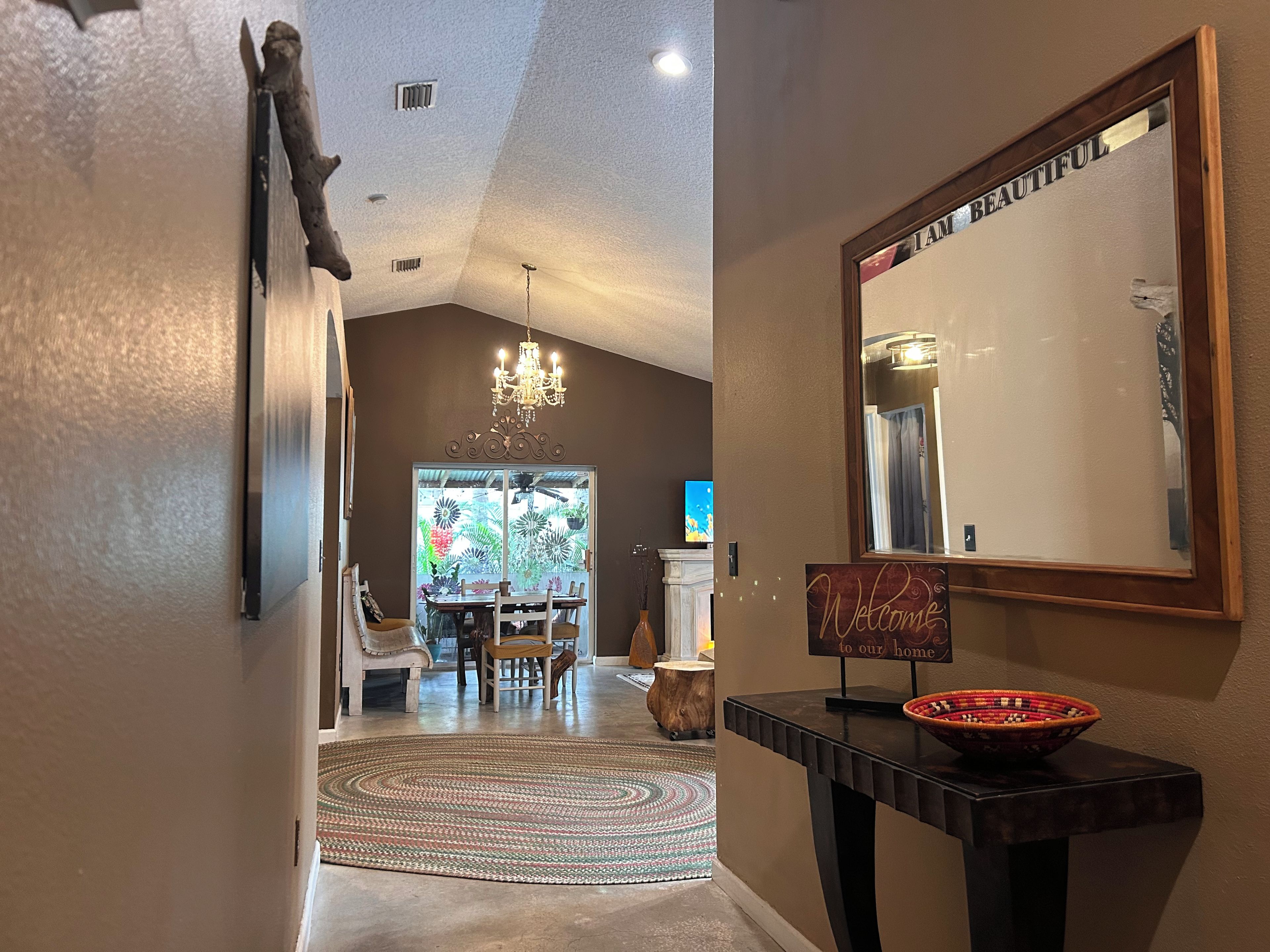 A hallway with a chandelier, a mirror, a welcome sign, and a dining area with chairs and a table.