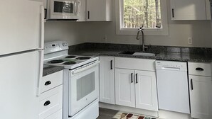 A kitchen with white cabinets, a stove, microwave, and a window with a view of trees. - Cozy cottage just one mile from the Platte river, close to the Sleepy Bear dunes (Honor)