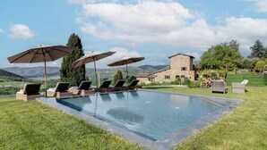 Una piscina con sillas de descanso y sombrillas, rodeada por un césped cuidado y un edificio de piedra al fondo.