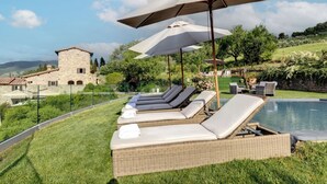 Zona de piscina con sillones y sombrillas, un edificio de piedra al fondo y cielo despejado.