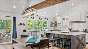 Une cuisine moderne dotée d’un îlot central, de poutres en bois, de luminaires suspendus et de grandes fenêtres.