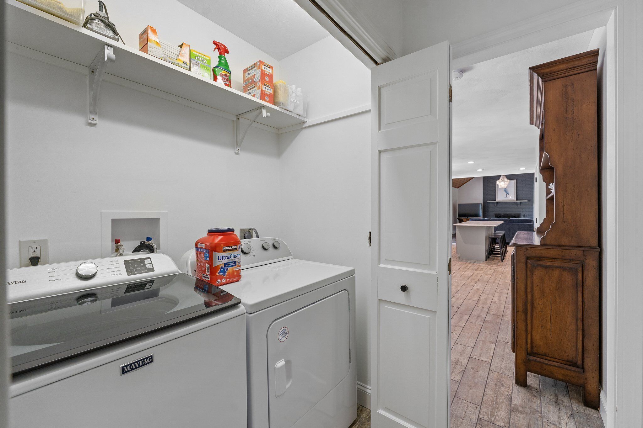 A laundry area with a front-loading washer and dryer, a shelf with cleaning supplies, and a wooden cabinet.