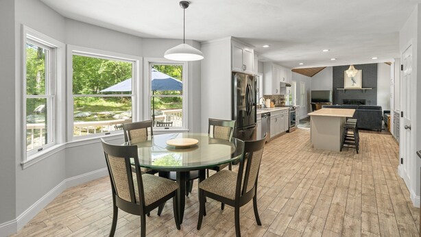A modern kitchen with a central island, large windows, and a dining area with a glass table. - Beautiful Colonial Home in Saunderstown, RI (North Kingstown)