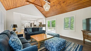 A spacious living room with a high ceiling, a wooden ceiling, a chandelier, a large sofa set, a coffee table, and a television. - Beautiful Colonial Home in Saunderstown, RI (North Kingstown)