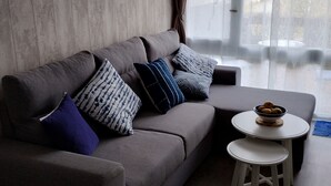A modern living room with a grey sofa, patterned cushions, a small round table with a bowl of fruit, and a view through large windows. - Cottage T2 (Lit-et-Mixe)