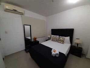 Quarto de hotel com cama, mesa de cabeceira, luminária, espelho e aparelho de ar condicionado.