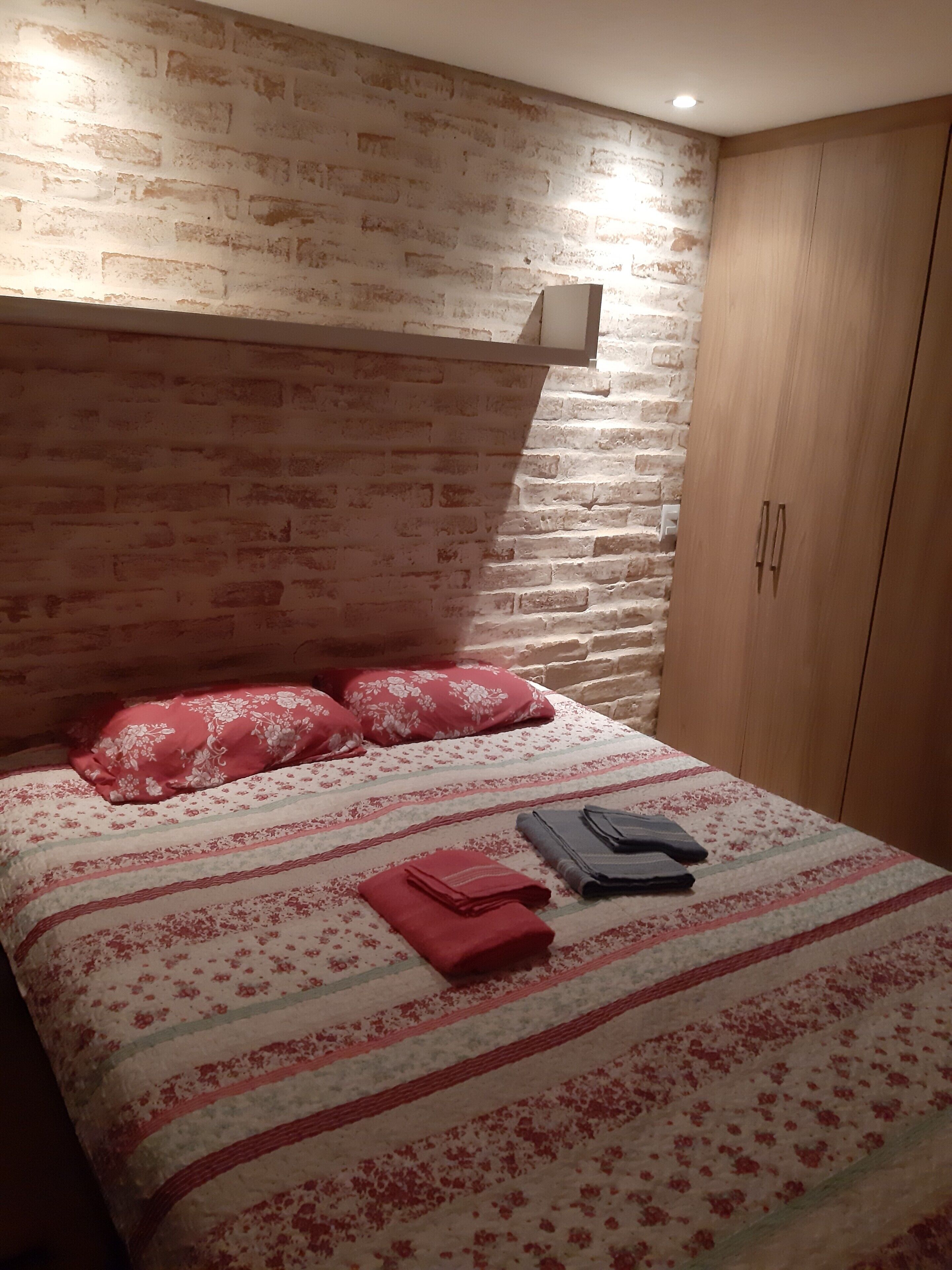Ein Schlafzimmer mit einem Kopfteil aus Backsteinoptik, einem Bett mit floraler Bettwäsche und einem Holzschrank.