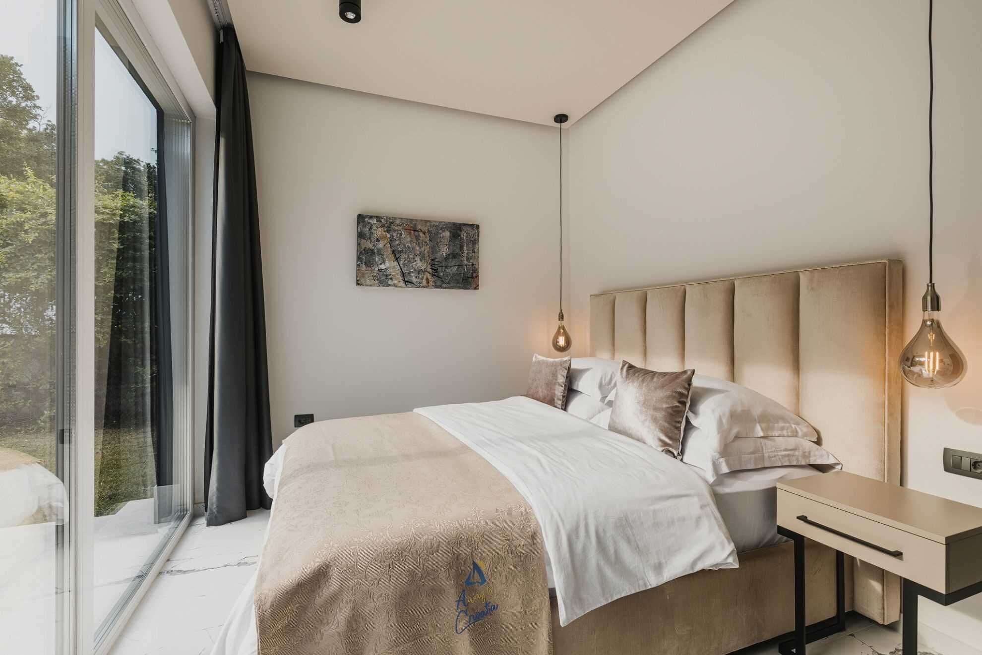 A modern bedroom with a large bed, bedside tables, a desk, and a large window.