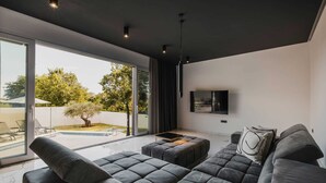 A modern living room with a large grey sectional sofa, a flat-screen TV mounted on the wall, and a glass door leading to a pool area. - Modern villa for 6 guests with pool, surrounded by pine forest, near beach (Valica)