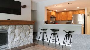 A modern kitchen with wooden cabinets, a built-in oven, a stainless steel refrigerator, and a stone fireplace.